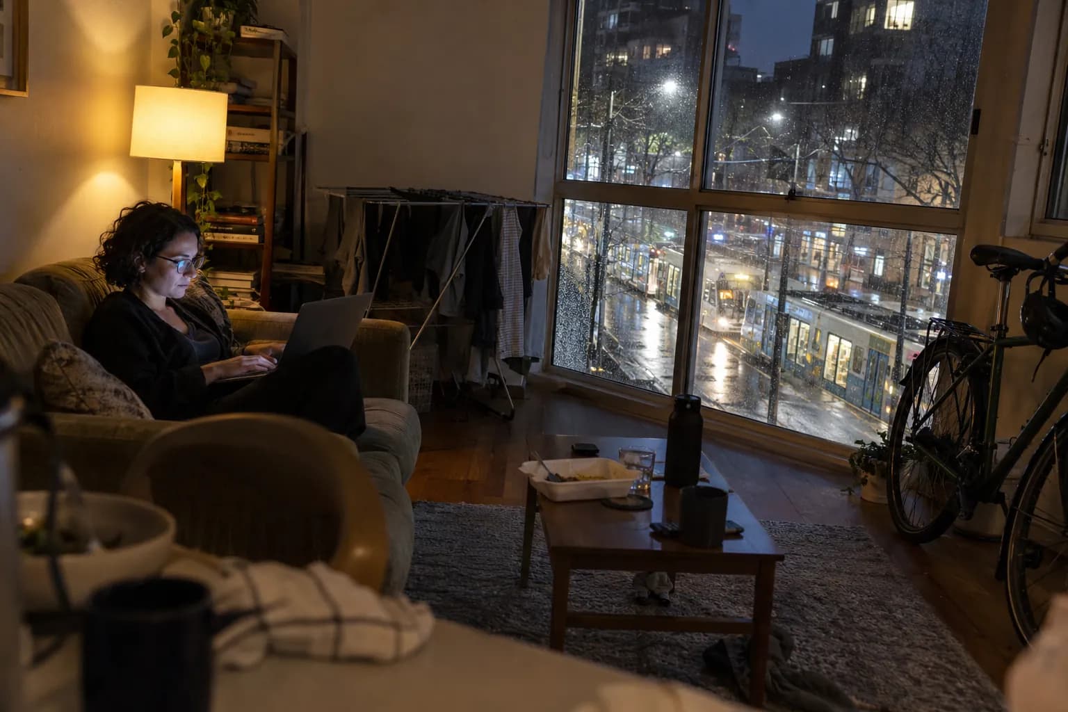 A woman on a couch with her laptop by a rainy apartment window