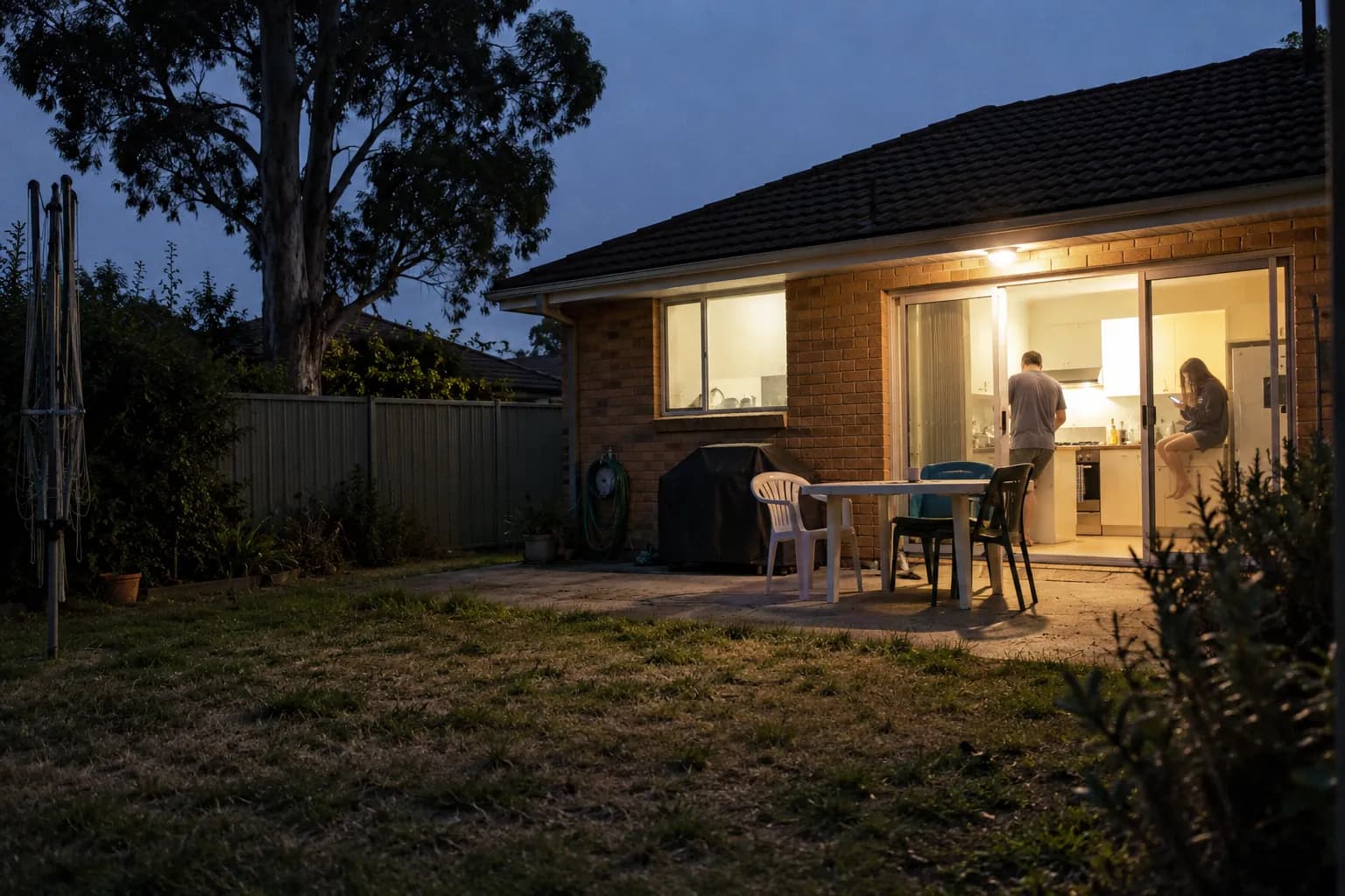 The back of an ordinary brick house at dusk: the kitchen light is on, a man is cooking and a teenager sits on the bench with her phone