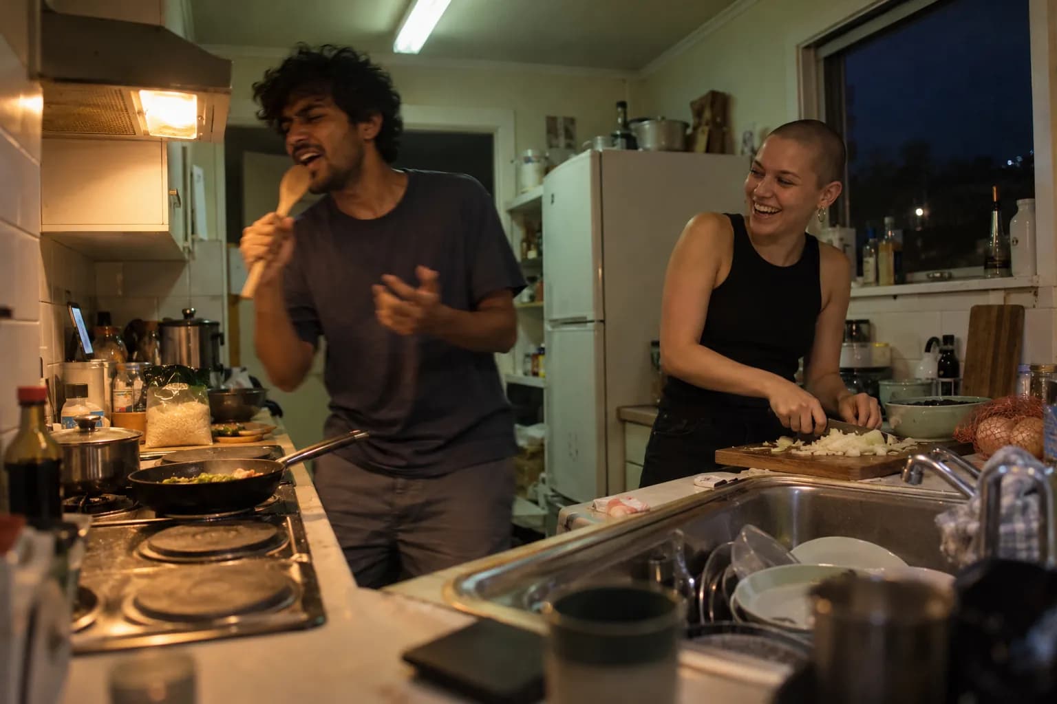 Two flatmates cooking at night, one singing into a wooden spoon