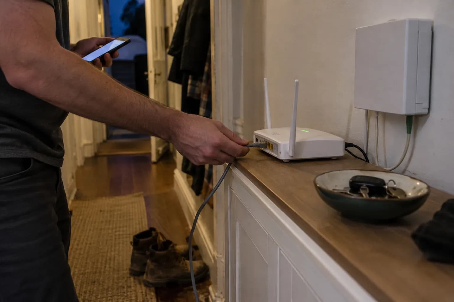 Someone plugging a network cable into a wi-fi router in their hallway, phone in the other hand